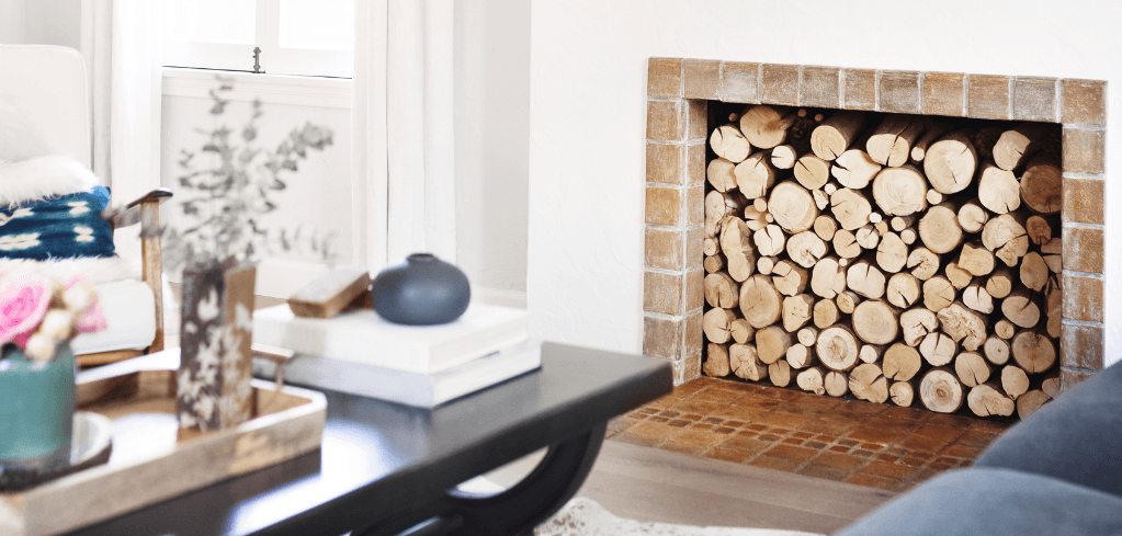 A cozy living room with a brick fireplace stacked with logs, a dark coffee table with decor, and a bright window nearby.
