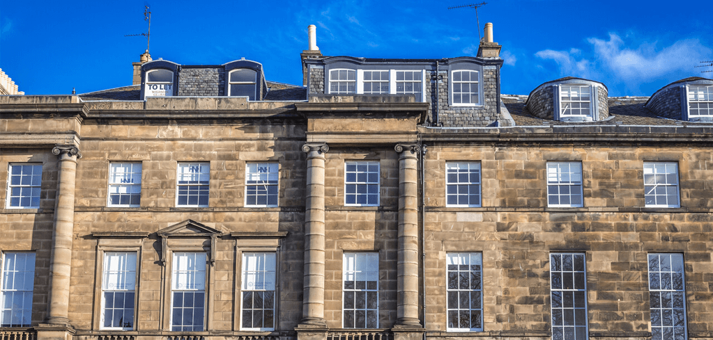 Historic stone townhouses with large windows against a bright blue sky, featuring a "TO LET" sign on the upper floor.