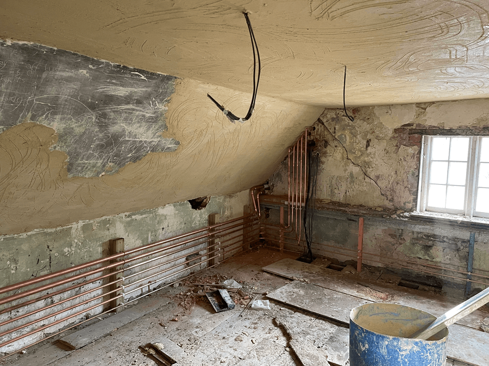 Dilapidated attic room under renovation with exposed copper pipes, crumbling plaster walls, and a window letting in light.