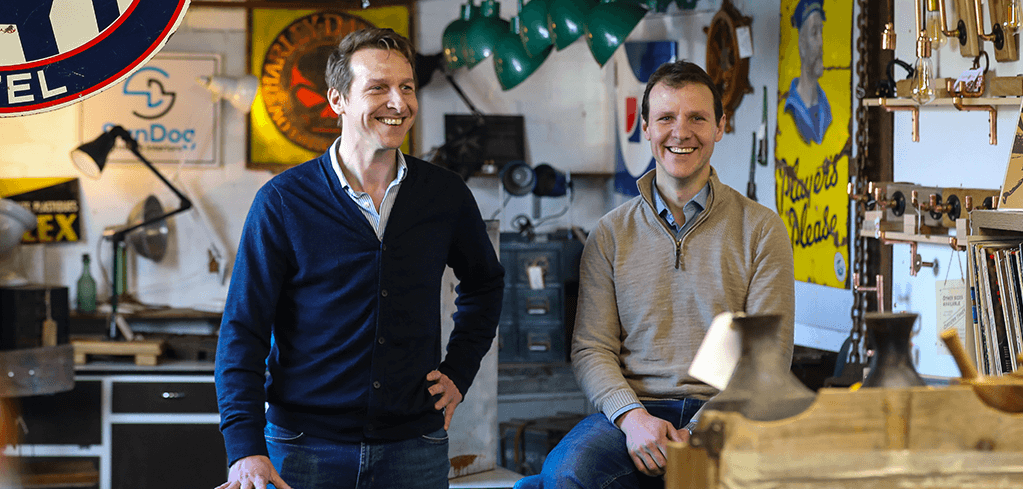 Two smiling men in sweaters standing in a workshop filled with vintage signs, tools, and collectibles.