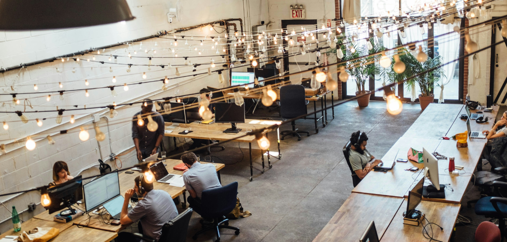 Modern coworking space with string lights, wooden desks, potted plants, and people working on laptops in an industrial setting.
