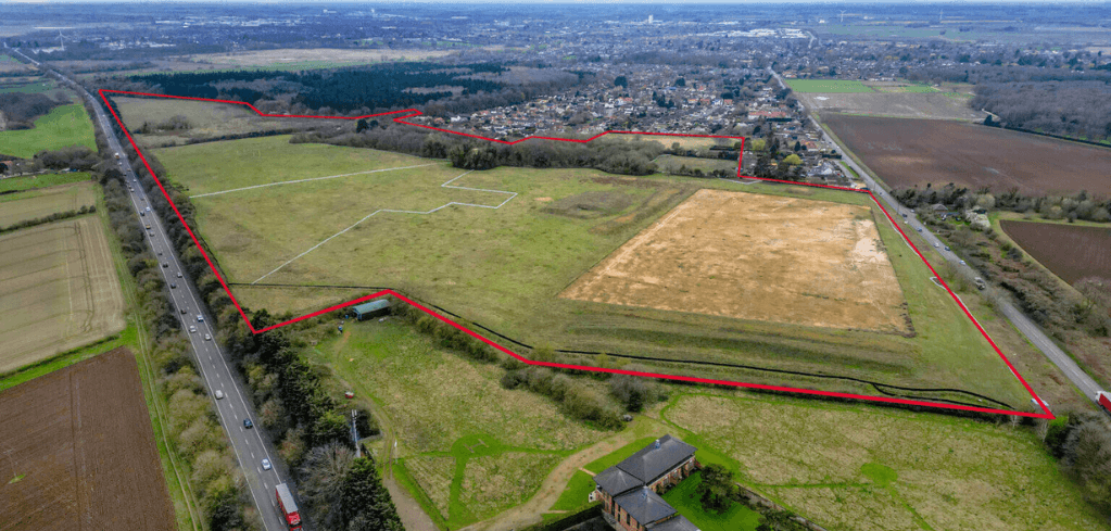 Aerial view of a large land parcel outlined in red, adjacent to a road and residential area, with green and brown fields.