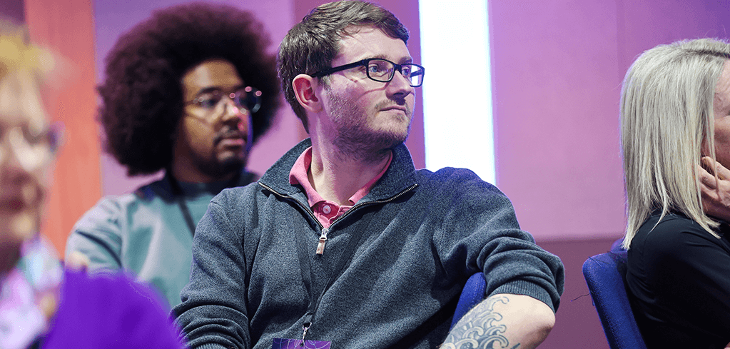 Diverse audience members seated in blue chairs at an event with purple lighting, focused on an unseen presenter.