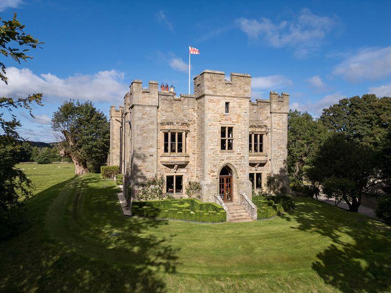 Historic stone castle with crenellated towers and flag flying, surrounded by manicured lawn and trees under blue sky.