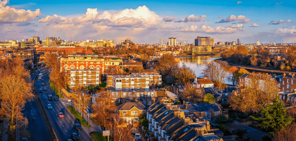 Aerial panoramic view of a London suburb at golden hour, featuring brick buildings, a busy road, a river, and a city skyline.