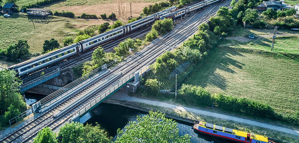 Aerial view of train tracks crossing a bridge over a canal with a colorful narrowboat below, surrounded by green countryside.