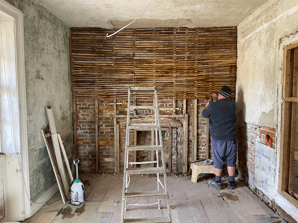 Person renovating old room with exposed brick fireplace, wooden lath wall, ladder, and worn plaster walls.