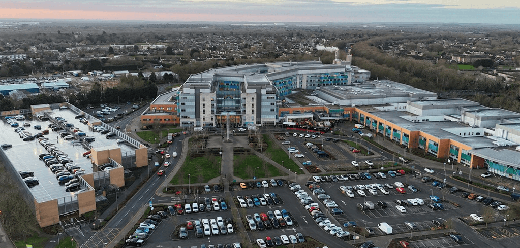 Aerial view of a large hospital complex with blue and orange buildings, surrounded by parking lots and suburban landscape.