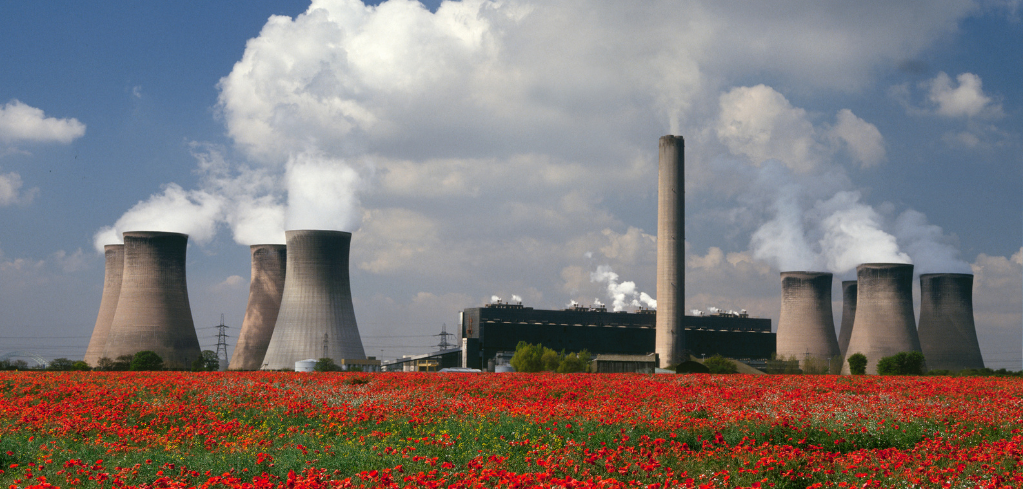 Power plant cooling towers with steam rising behind a vibrant field of red poppies under a blue, cloudy sky.