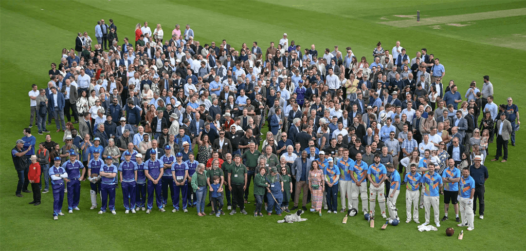 Large crowd gathered on a cricket field, including players in blue uniforms and spectators in formal and casual attire.
