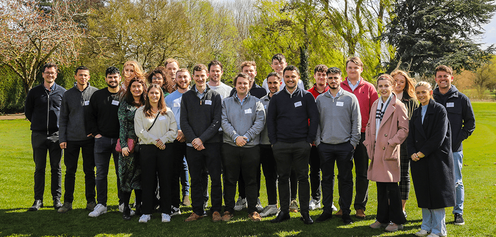 Group of young professionals wearing name badges gathered outdoors on green lawn with trees in background on sunny day.
