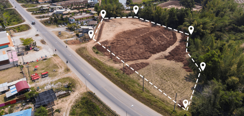 Aerial view of a vacant land plot outlined with a dashed white line and location pins, near a road and residential area.