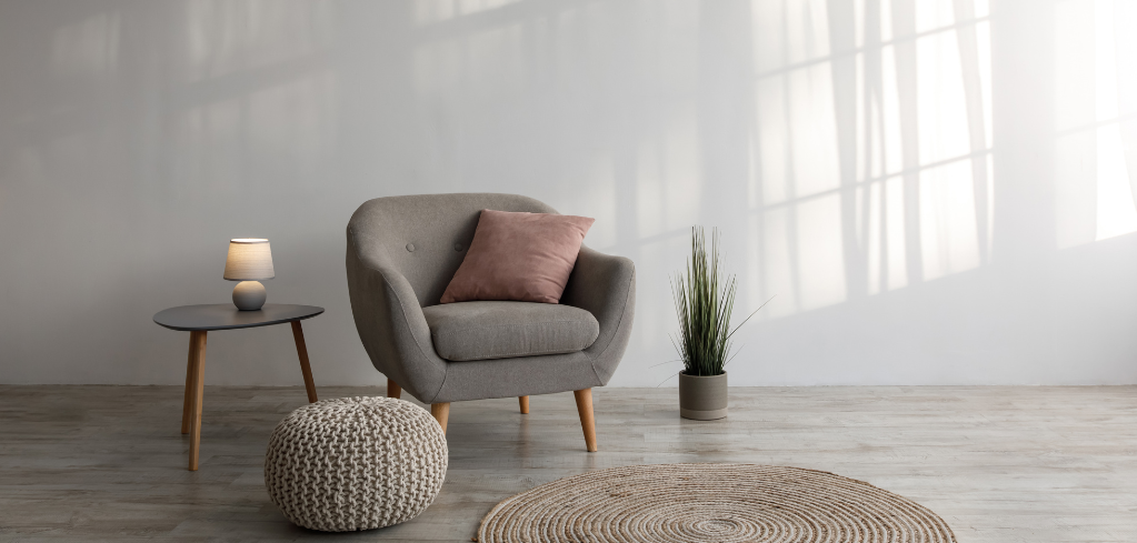 A cozy living space with a gray armchair, pink pillow, side table with lamp, knit pouf, round rug, and a potted plant.