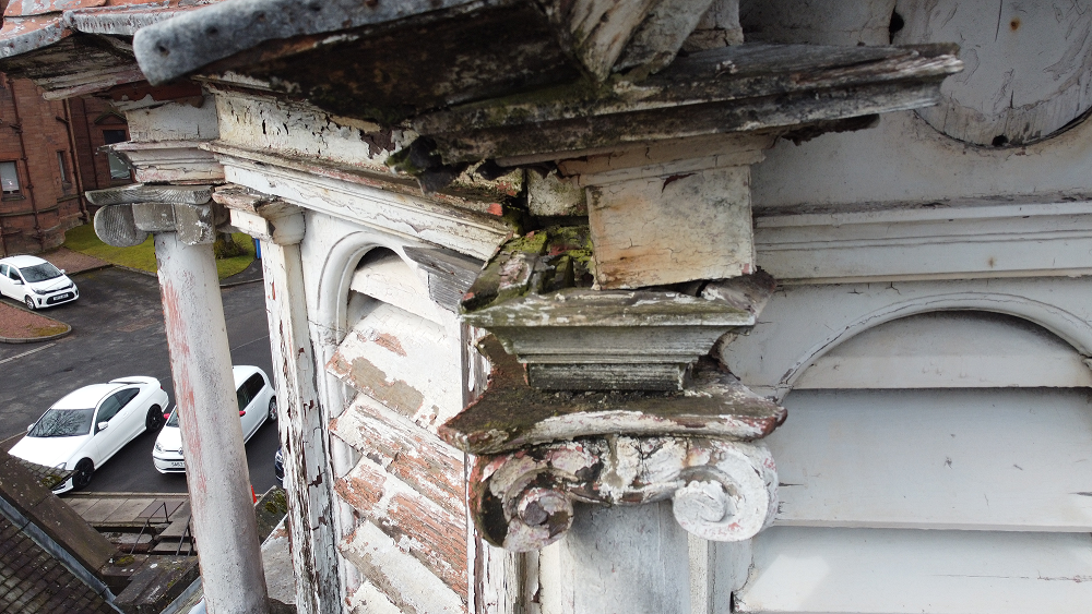Severely deteriorated architectural detail on a building with peeling paint and rotting wood, overlooking a parking area.