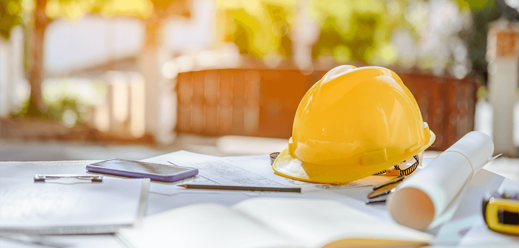 Yellow hard hat on construction blueprints with smartphone and rolled plans at outdoor worksite.