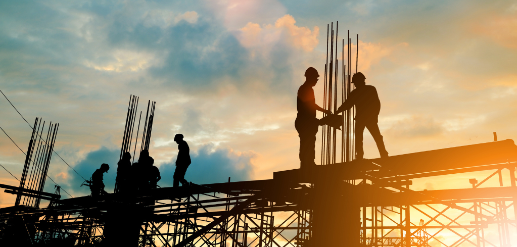 Silhouettes of construction workers on a building framework against a vibrant orange and blue sunset sky.