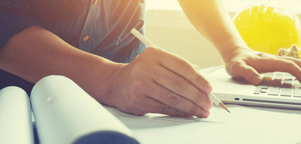Person writing notes with pencil while working on laptop in warm sunlight, with rolled blueprints nearby.
