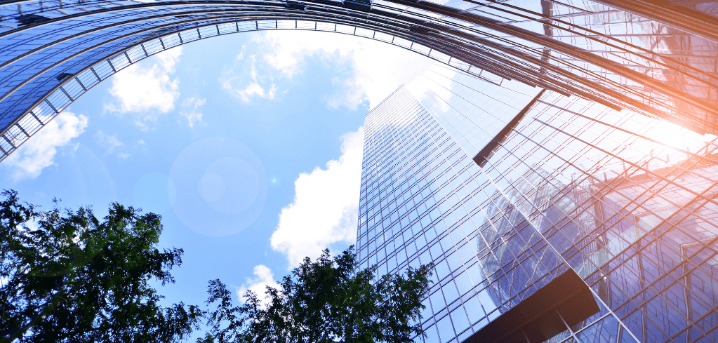 Upward view of tall glass skyscrapers against a blue sky with white clouds, green trees visible at the bottom left.