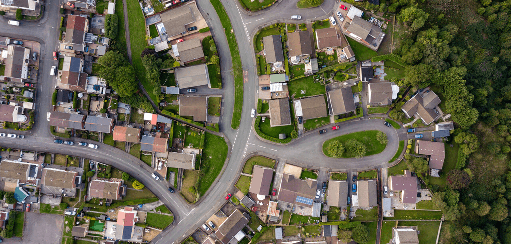 Aerial view of a suburban neighborhood with winding roads, houses with brown roofs, green lawns, and bordering forest area.