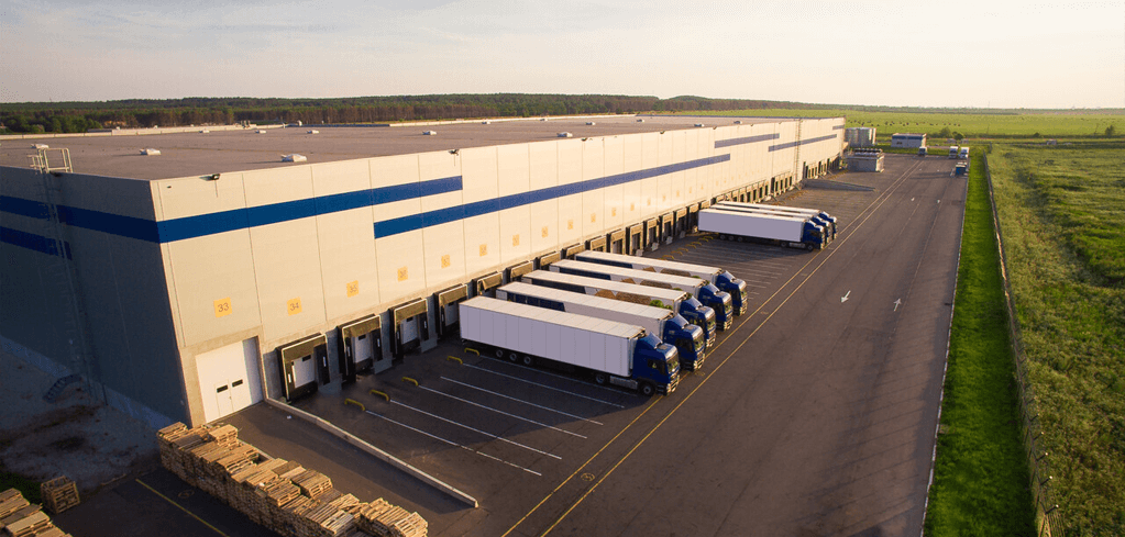 Large distribution warehouse with blue and white exterior, loading docks, and several semi-trucks parked alongside green fields.
