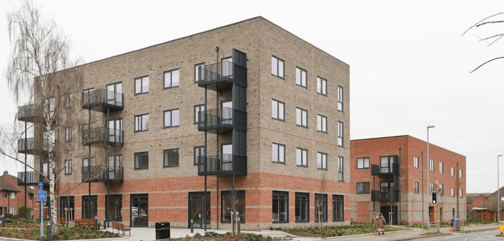 Modern four-story brick apartment building with balconies, ground-floor retail units, and a smaller adjacent building on a street corner.