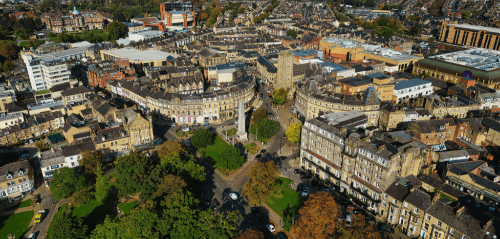 Aerial view of a historic English town with stone buildings, a central monument, tree-lined streets, and autumn foliage.