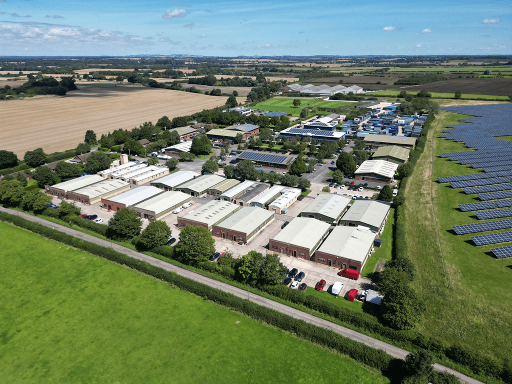 Aerial view of industrial park with warehouses surrounded by farmland, solar panels, and green fields under blue sky.