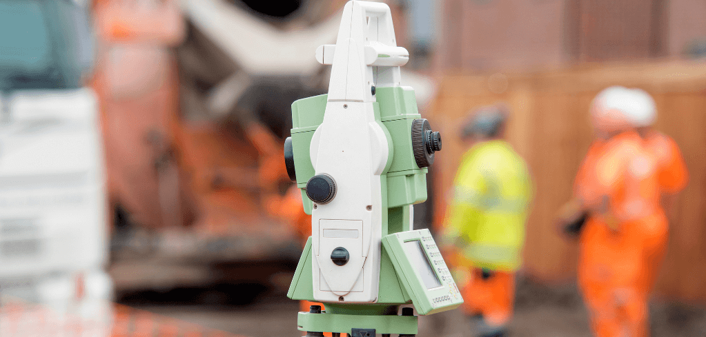 Green and white surveying equipment at a construction site with workers in orange safety vests in the background.