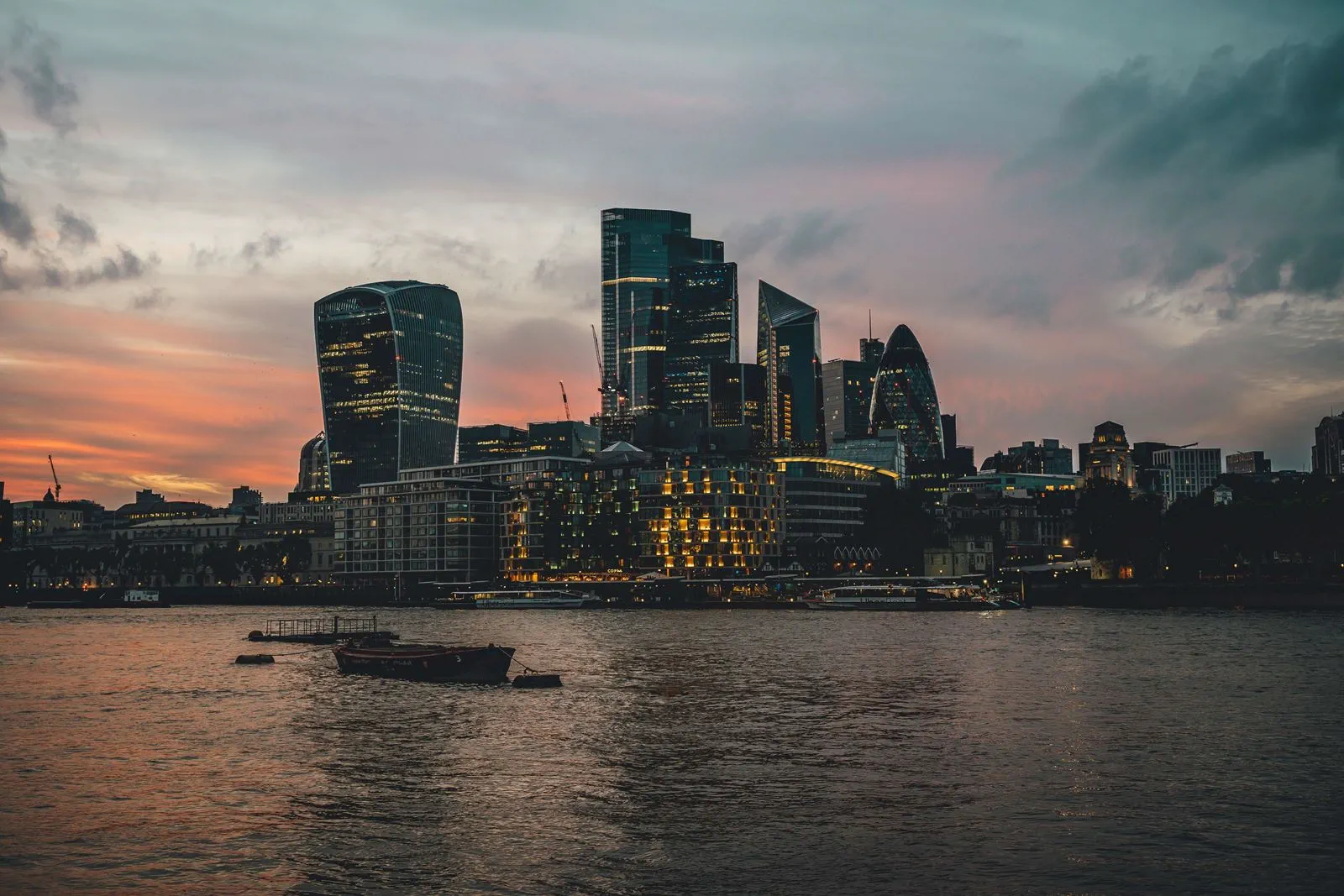 London skyline at sunset with illuminated skyscrapers including the Gherkin and Walkie Talkie buildings along the Thames River.