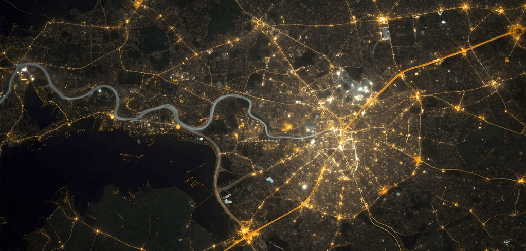 Aerial night view of a city with illuminated streets and a winding river, showing urban infrastructure from space.