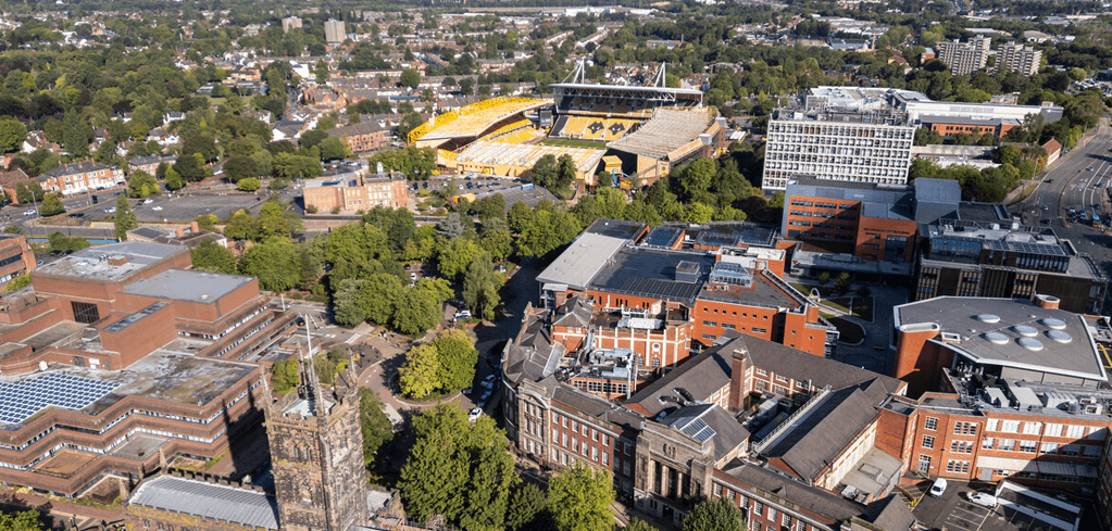 Aerial view of a city with a large yellow-seated sports stadium surrounded by university buildings, trees, and urban development.