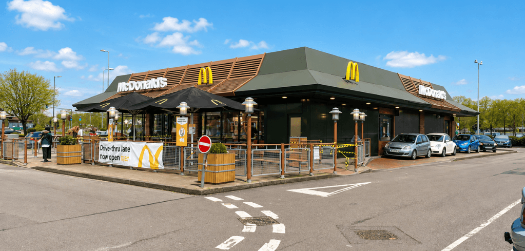 Exterior of a McDonald's restaurant with a dark roof, golden arches, outdoor seating, and a drive-thru lane open sign.