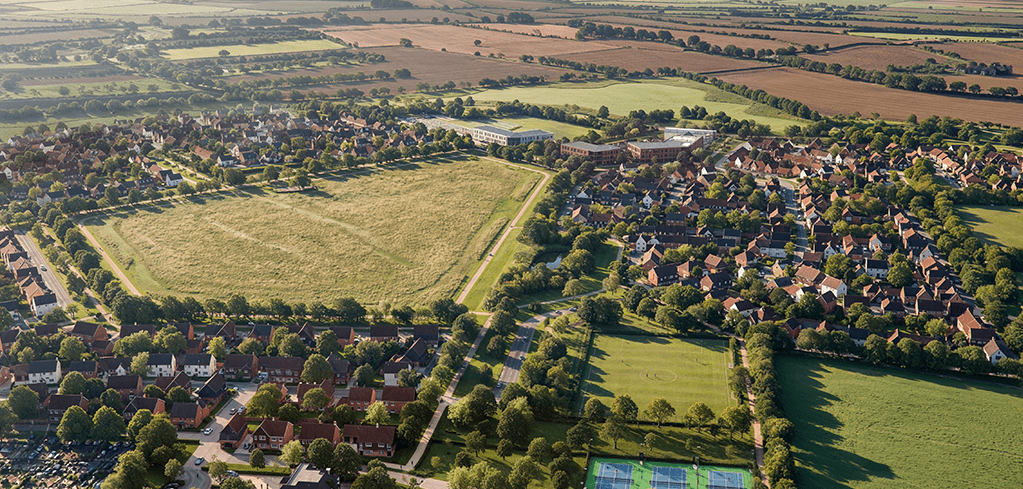 Aerial view of a English village with red-brick homes, green parks, tennis courts, and surrounding farmland fields.