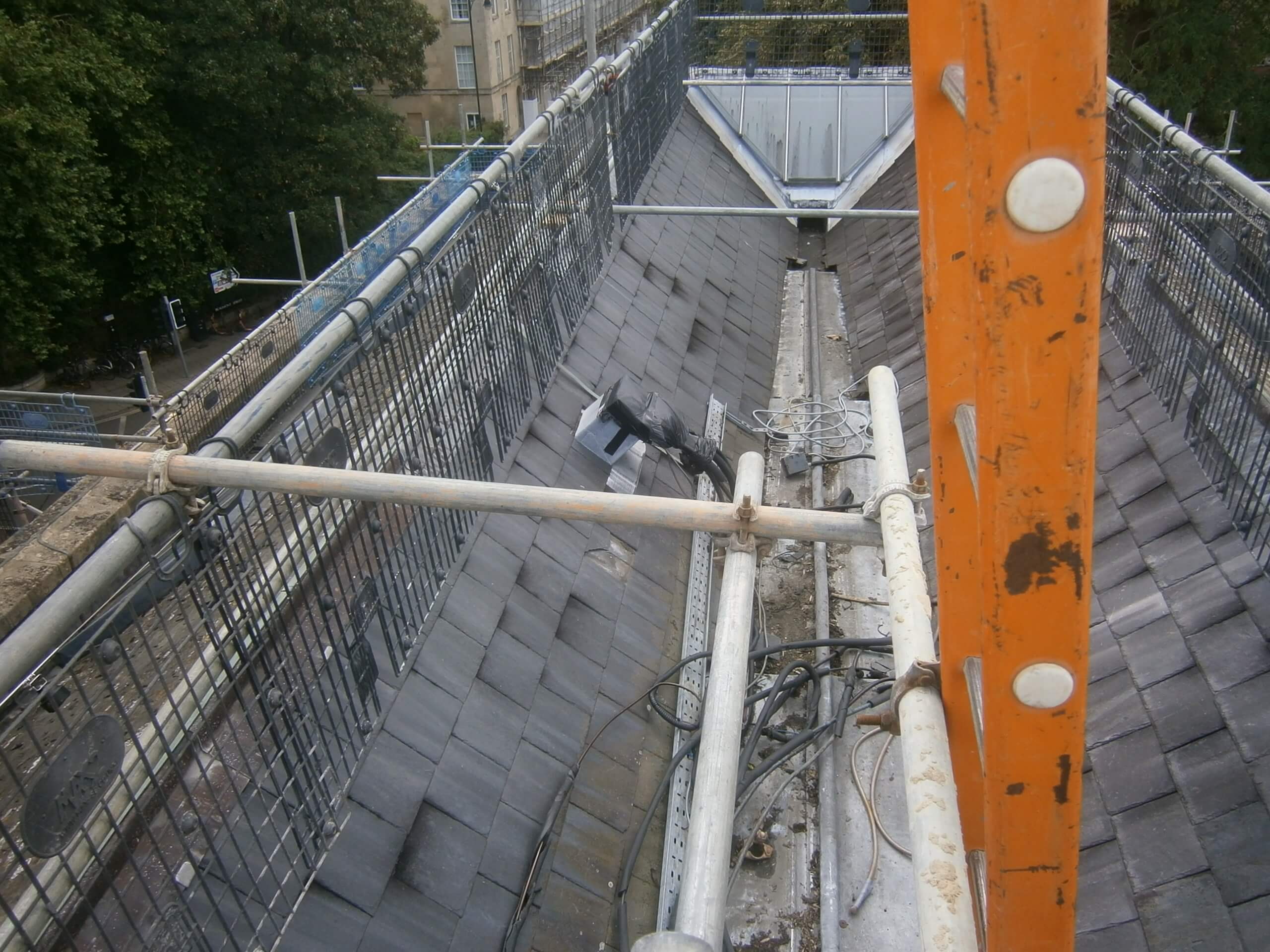 Aerial view of a slate roof under construction with metal scaffolding, orange support beam, cables, and mesh safety barriers.