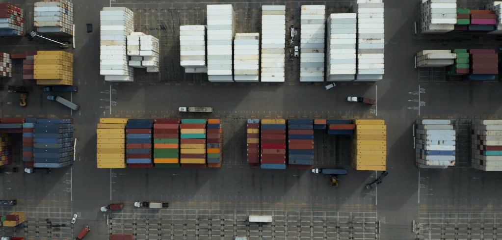 Aerial view of a shipping container yard with colorful stacked containers and trucks moving through organized lanes.