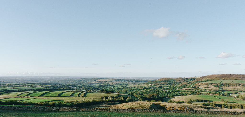 Panoramic view of rolling green countryside with patchwork fields, gentle hills, and a clear blue sky with scattered clouds.
