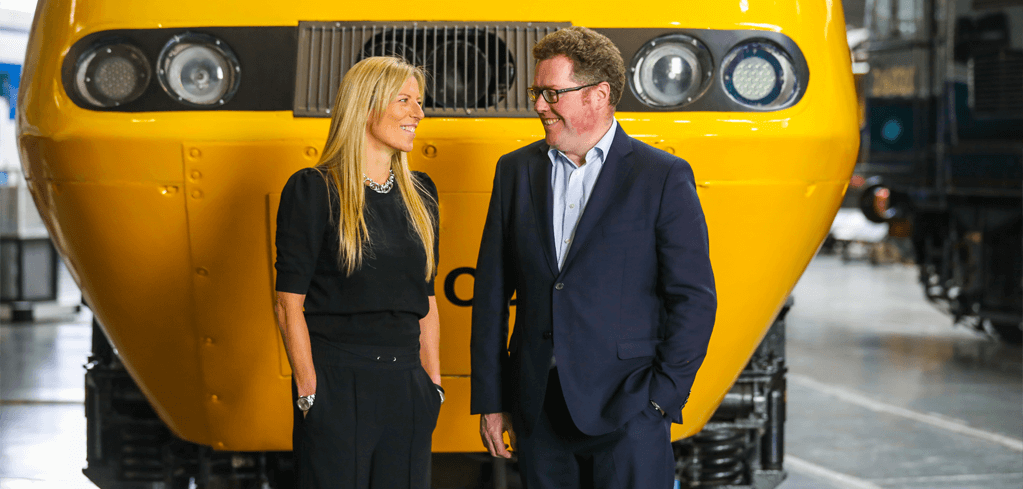 Two professionals in business attire smiling at each other in front of a bright yellow train or locomotive.