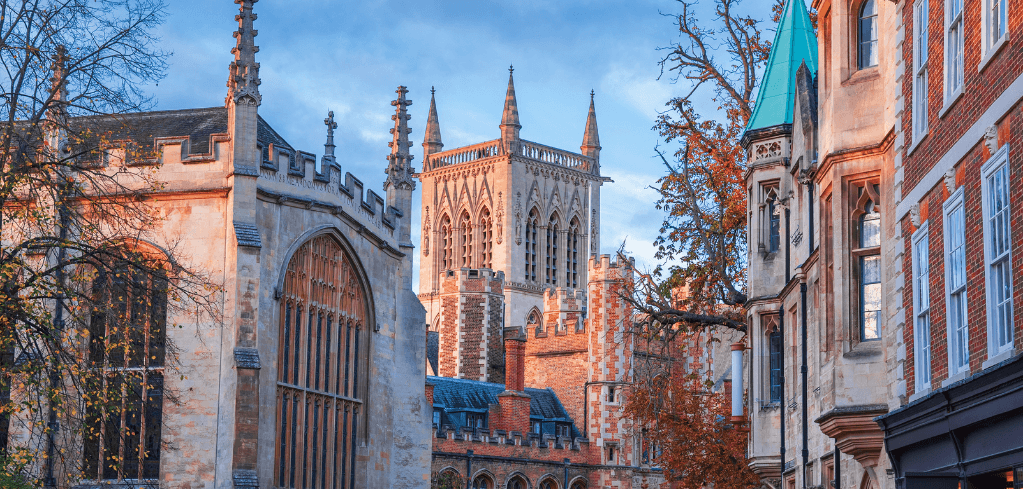 Gothic stone cathedral with spires rises between historic brick buildings and autumn trees under a blue sky.