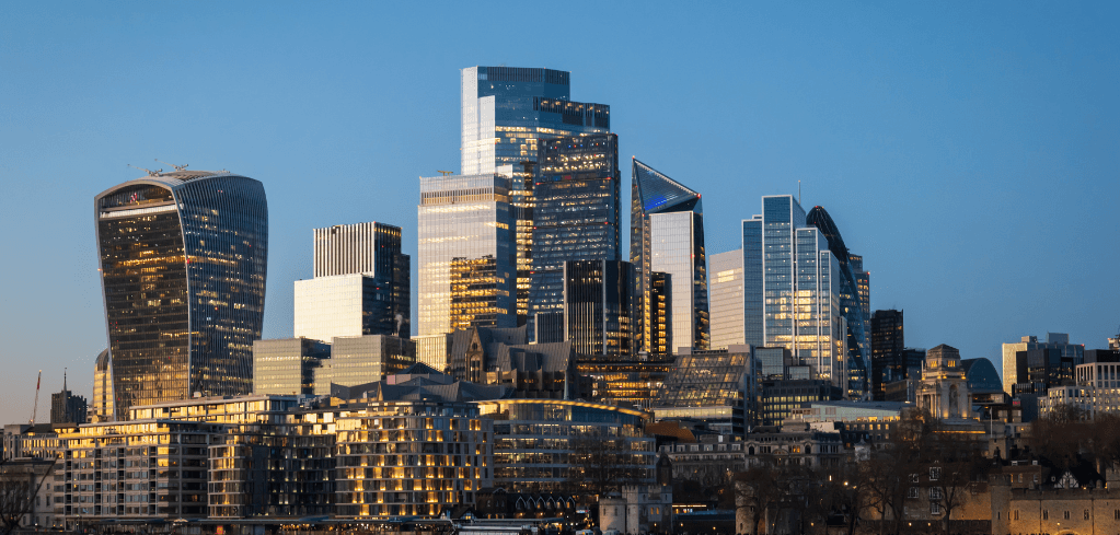 London's financial district skyline at dusk, featuring iconic glass skyscrapers lit with golden lights against a clear blue sky.