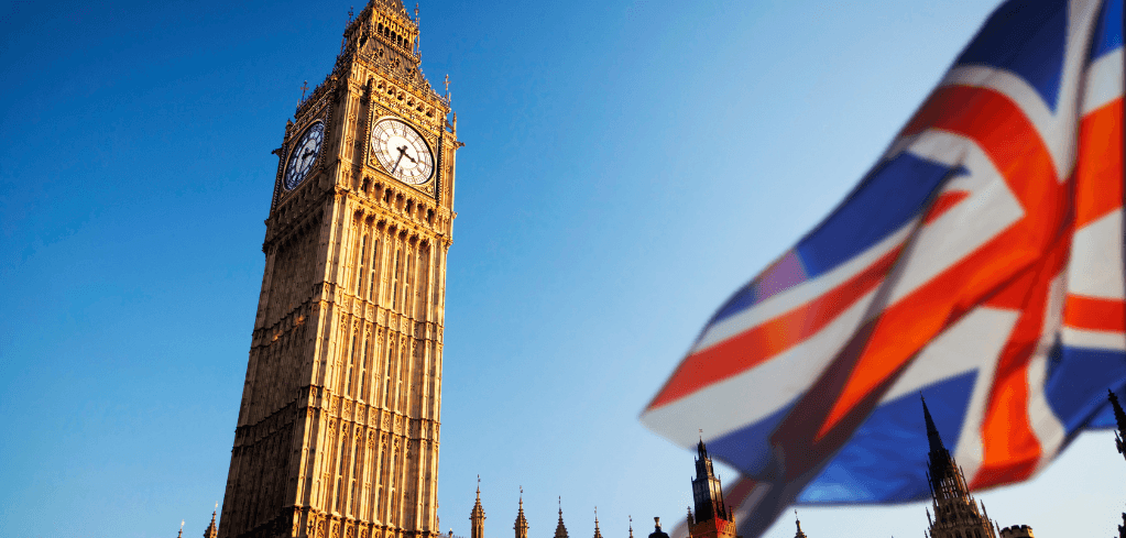 Big Ben clock tower in London against a blue sky, with a blurred Union Jack flag waving in the foreground.