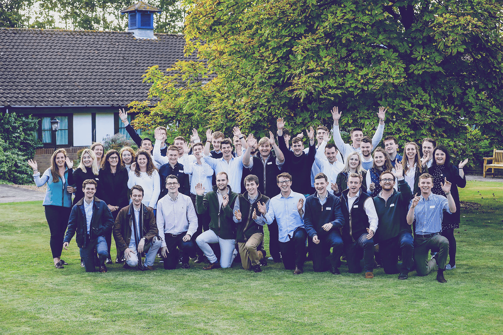 Large group of young professionals posing cheerfully with raised hands on a lawn in front of a building with lush trees.