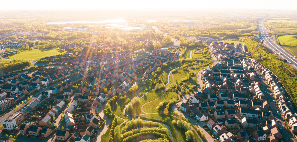 Aerial view of suburban housing development at sunset with winding green spaces, lakes, and highway in the distance.