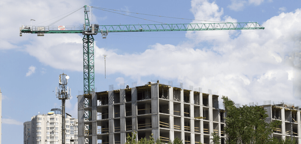 Green tower crane extending over a concrete building under construction, with blue sky and white clouds in background.