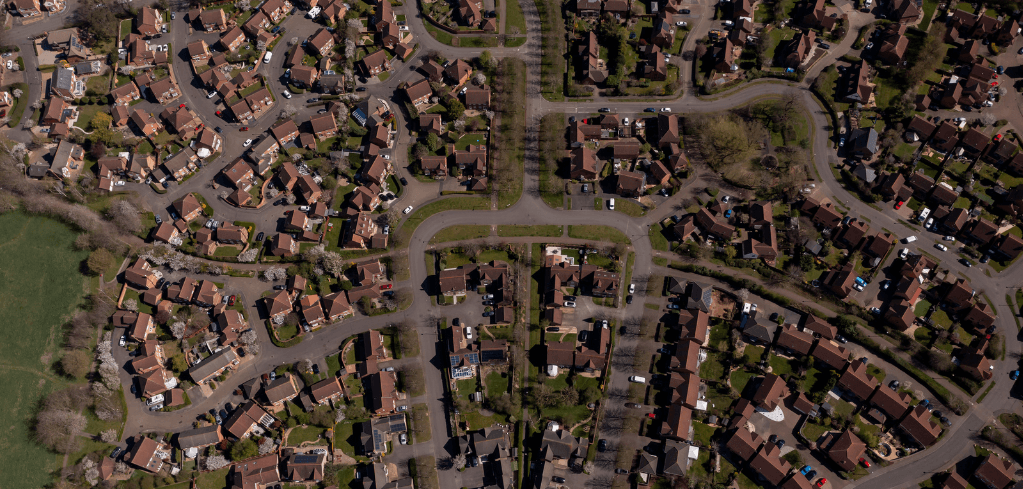 Aerial view of a residential neighborhood with brown-roofed houses, winding streets, green lawns, and parked cars.