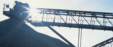 An industrial conveyor system transporting aggregate material, with sunlight shining through the metal framework and a gravel mound below.
