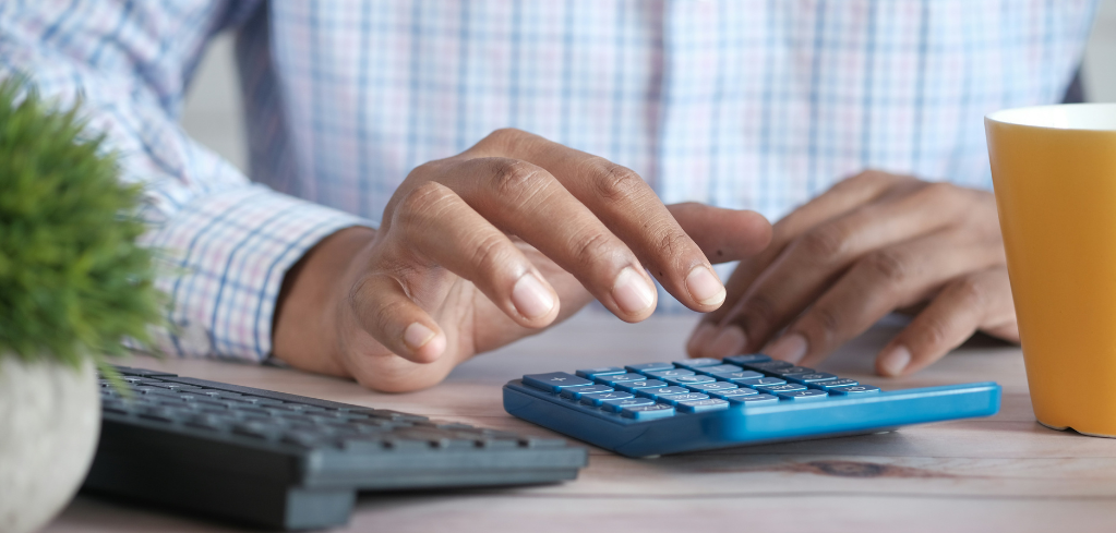 Person in plaid shirt using blue calculator at desk with small plant and yellow mug nearby.