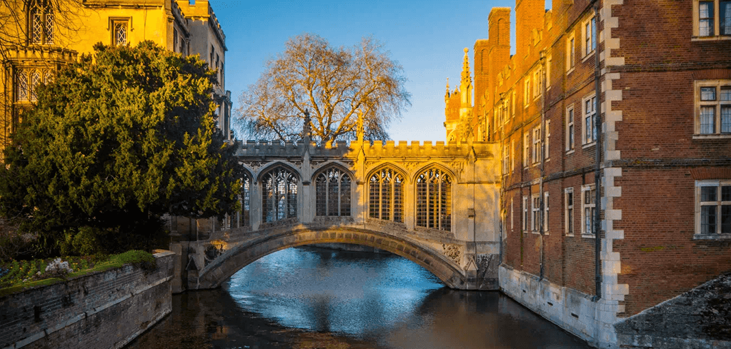 Historic Bridge of Sighs at Cambridge University bathed in golden sunset light, spanning a narrow canal between brick buildings.