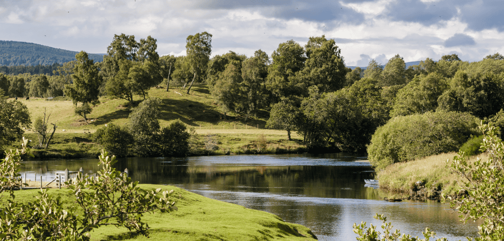 Serene countryside landscape with a calm river winding through green hills and trees under a cloudy sky.
