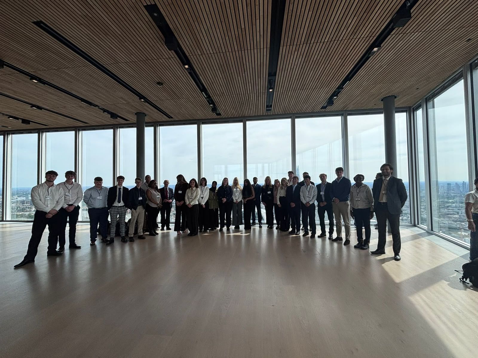 A large group of professionally dressed people posing in a modern high-rise room with floor-to-ceiling windows overlooking a city skyline.