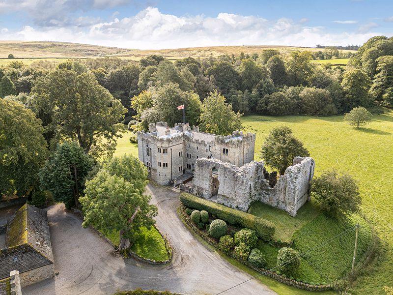 Aerial view of a historic stone castle with partially ruined walls, surrounded by lush green countryside and forests.